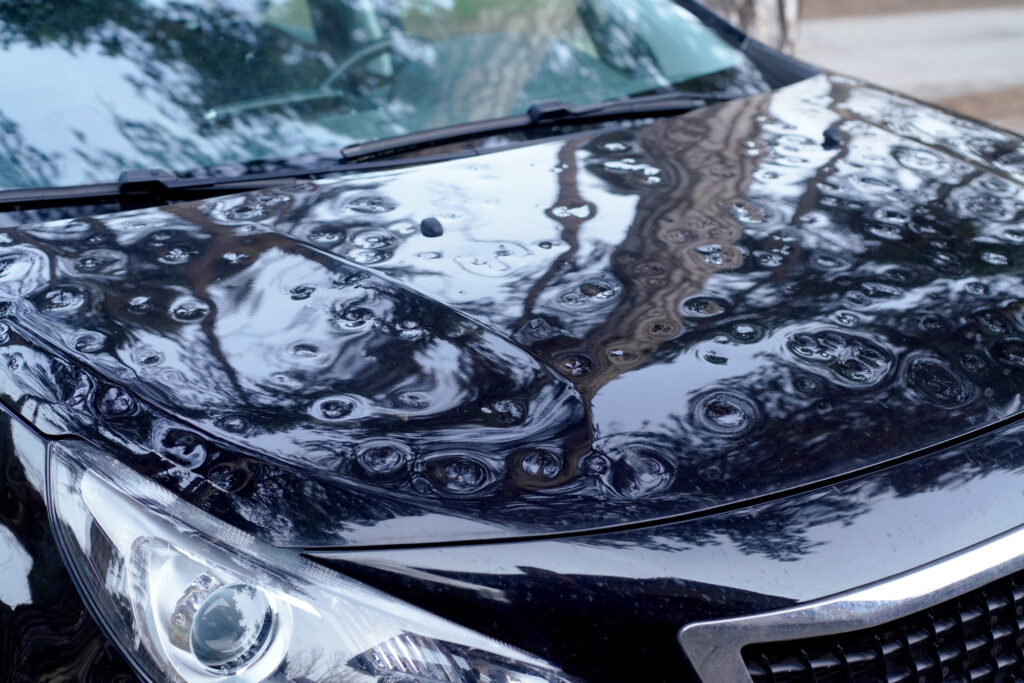 Close-up of a black car hood covered in extensive hail damage dents after a storm, showing circular impact marks across the surface.